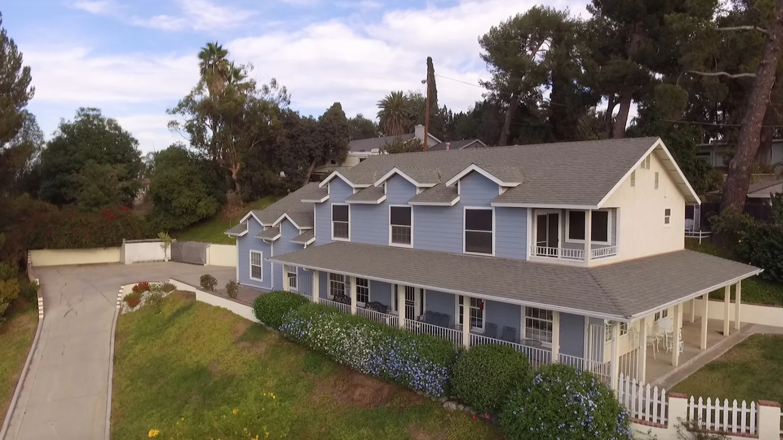 A two-story blue farmhouse-style home on a Riverside hillside lot, wraparound covered porch, white picket fence and a flowering hedge along the slope